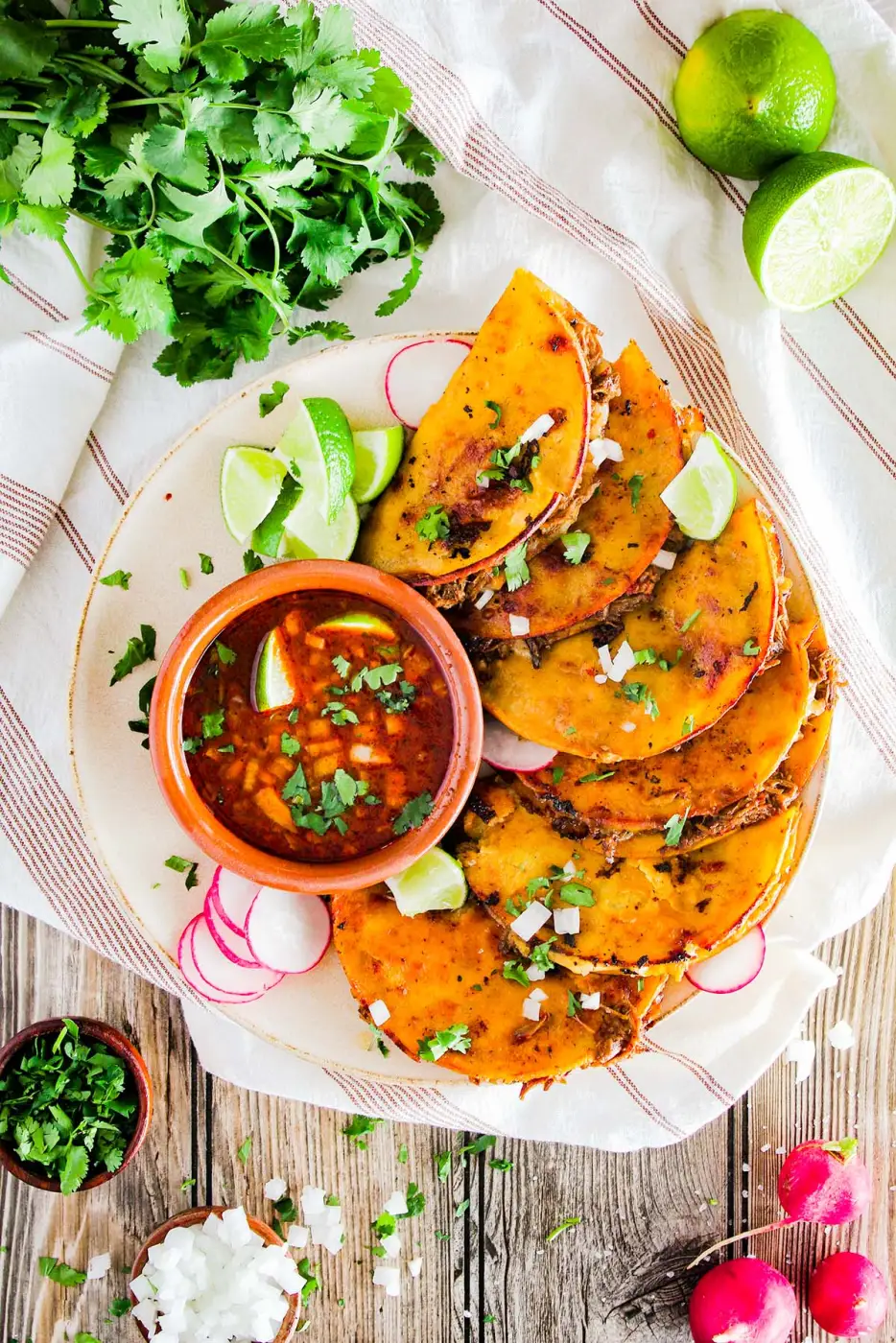 Overhead birria taco spread with consommé and garnishes